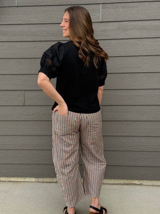 Woman wearing a black top and striped pants standing against a gray wooden wall.