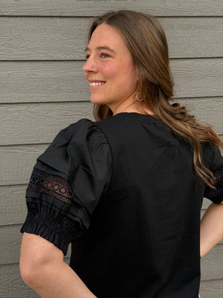 Woman wearing a black blouse with lace sleeves against a gray wooden wall.