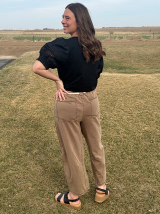 Woman standing outdoors in a field wearing a black top and beige pants.