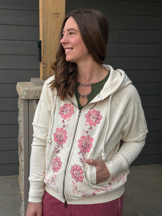 Woman wearing a cream-colored hoodie with pink floral patterns, standing against a dark wooden door.