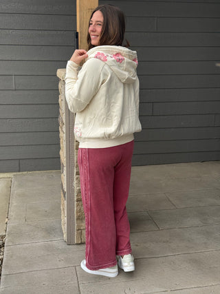 Person wearing a white hoodie with floral designs and pink pants standing against a gray wall.