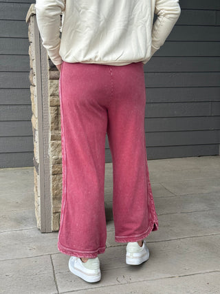 Person wearing pink wide-leg pants and a cream jacket standing against a wooden wall.