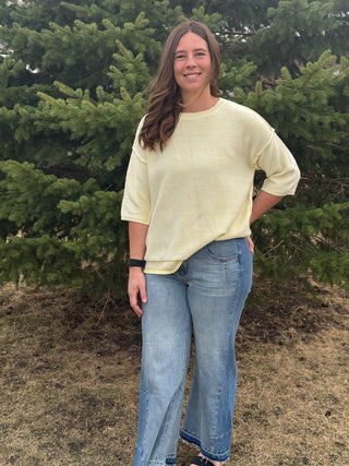 Woman wearing a light yellow shirt and blue jeans standing in front of green trees.