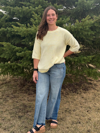 Woman wearing a light yellow sweater and blue jeans standing in front of green trees.