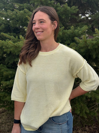 Woman wearing a light yellow sweater standing in front of green trees
