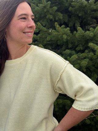 Woman wearing a light yellow sweater standing in front of green trees