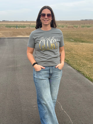 Woman wearing a gray t-shirt with 'God gets the Glory' text, standing on a road with fields in the background.
