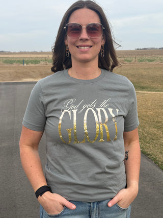 Woman wearing a gray t-shirt with 'God gets the GLORY' printed on it, standing outdoors.