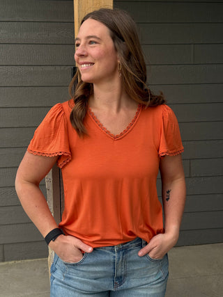 Woman wearing an orange top and blue jeans standing against a gray wall.