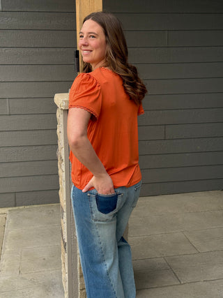 Woman in an orange shirt and blue jeans standing against a gray wall.