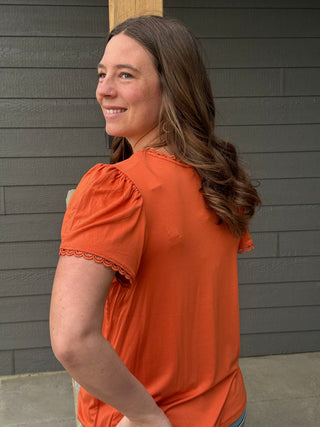 Woman wearing an orange blouse standing against a gray wooden wall.