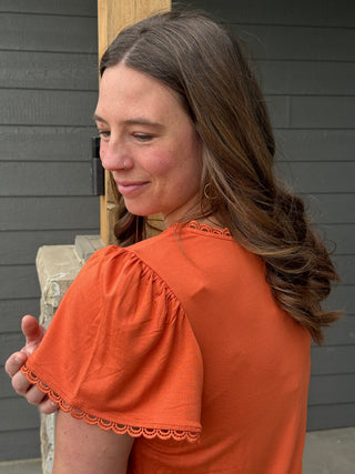 Woman wearing an orange blouse with lace details, standing against a gray wall.