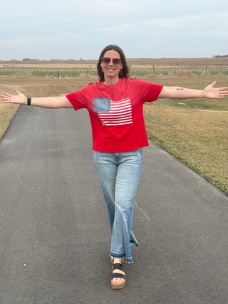 Person wearing a red t-shirt with an American flag design, standing on a road with open arms.