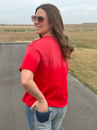 Woman wearing a red shirt and sunglasses, standing on a road with fields in the background