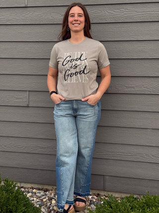 Woman wearing a gray t-shirt with text and blue jeans standing against a gray wall.