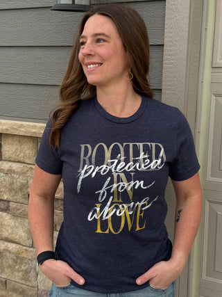 A lady wearing a blue graphic tee with "Rooted in Love, Protected from above" standing by a house.