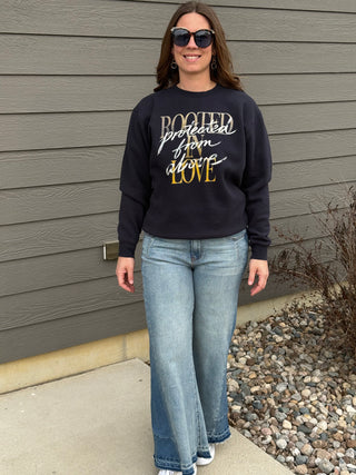 Woman wearing a black sweatshirt with text and blue jeans standing against a gray wall.