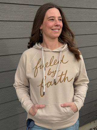 Woman wearing a beige hoodie with 'fueled by faith' text against a gray wall.