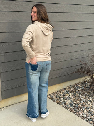 Woman wearing a beige hoodie and blue jeans standing against a gray wall.