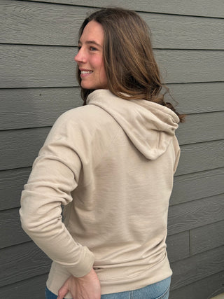 Woman wearing a beige hoodie standing against a gray wall.
