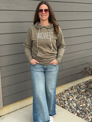 Woman wearing a green hoodie with 'Midwest' text, blue jeans, and sunglasses standing against a gray wall.