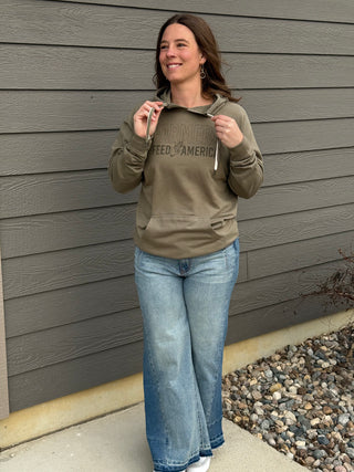 Woman wearing a green hoodie and blue jeans standing against a gray wall.