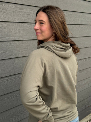 Woman wearing a beige hoodie standing against a gray wall.