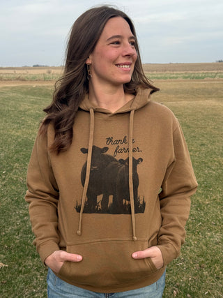 Woman wearing a brown hoodie with cow design and 'thank a farmer' text in a field.