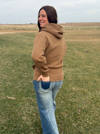 Woman wearing a brown hoodie and blue jeans standing in an open field.