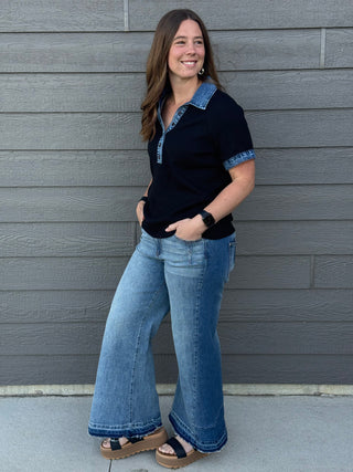 Woman wearing a black shirt with blue trim and blue jeans standing against a gray wall.