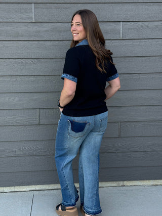 Woman wearing a black shirt with blue accents and blue jeans standing against a gray wall.