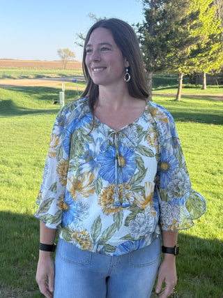 Woman wearing a floral blouse standing in a grassy field with trees in the background