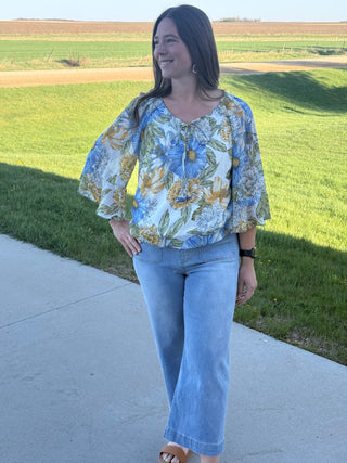 Woman wearing a floral blouse and jeans standing on a sidewalk with a grassy field in the background