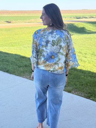 Woman wearing a floral blouse and blue jeans standing on a sidewalk with a field in the background