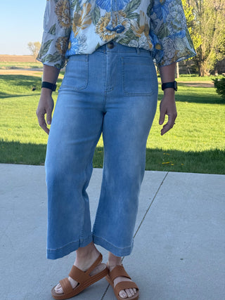 Person wearing a floral top and blue jeans standing on a sidewalk with grass and trees in the background.