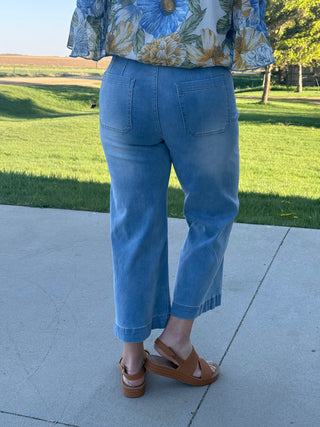 Person wearing a floral top and blue jeans standing on a sidewalk with grass and trees in the background.