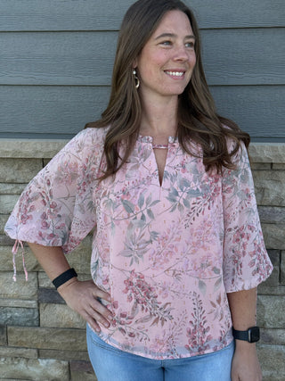 Woman wearing a pink floral blouse standing against a stone wall.