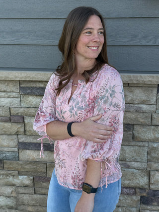 Woman wearing a pink floral blouse and blue jeans standing against a stone wall.
