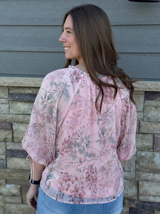 Woman wearing a pink floral blouse standing in front of a stone wall.