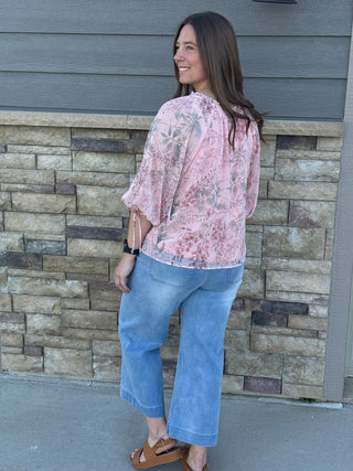Woman wearing a pink floral blouse and blue jeans standing against a stone wall.