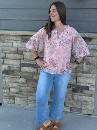 Woman wearing a floral blouse and blue jeans standing against a stone wall.