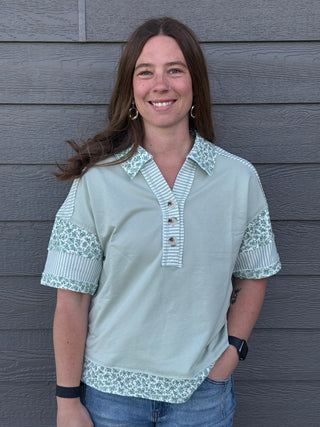 Woman wearing a light green blouse with floral patterns against a gray wooden wall.