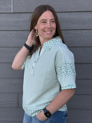 Woman wearing a light blue shirt with floral patterns against a gray wooden wall.