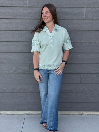 Woman wearing a light green blouse with lace trim and blue jeans standing against a gray wooden wall.