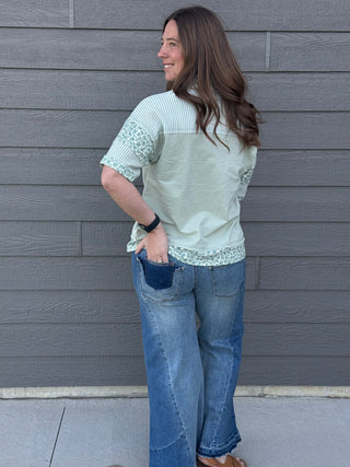 Woman wearing a light green and white patterned top with blue jeans against a gray wall.