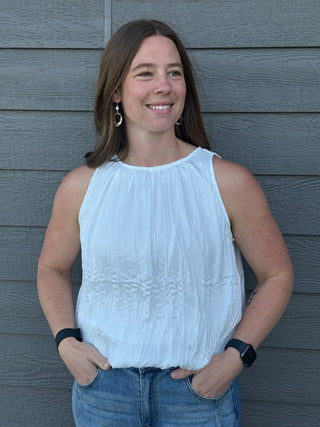 Woman wearing a white sleeveless top and blue jeans standing against a gray wooden wall.