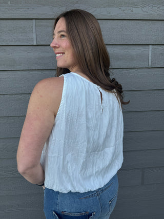 Woman wearing a white sleeveless top and blue jeans against a gray wooden panel background