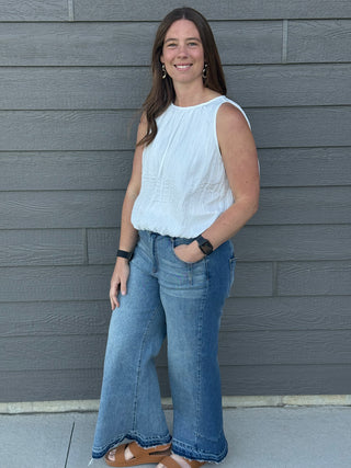 Woman wearing a light blue sleeveless top and blue jeans standing against a gray wall.
