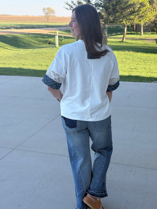 Person wearing a white shirt with blue accents and jeans, standing on a concrete surface with grass and trees in the background.