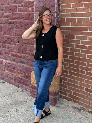A lady wearing a black sweater vest with big gold buttons against a brick wall.